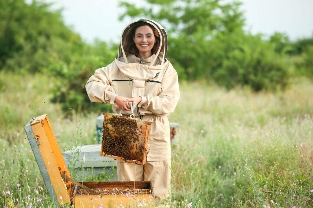 Eine Imkerin in Schutzkleidung hält einen mit Bienen besetzten Bienenstockrahmen auf einer Blumenwiese, um Propolis zu ernten