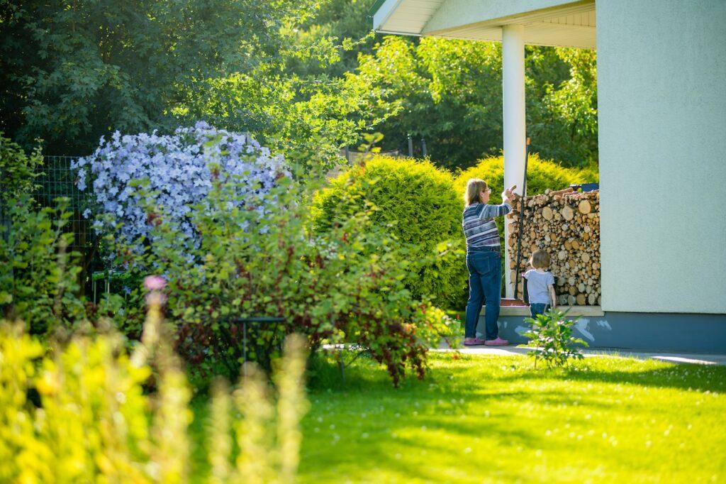 Frau und Kind stapeln Feuerholz neben einem Haus im gruenen Garten