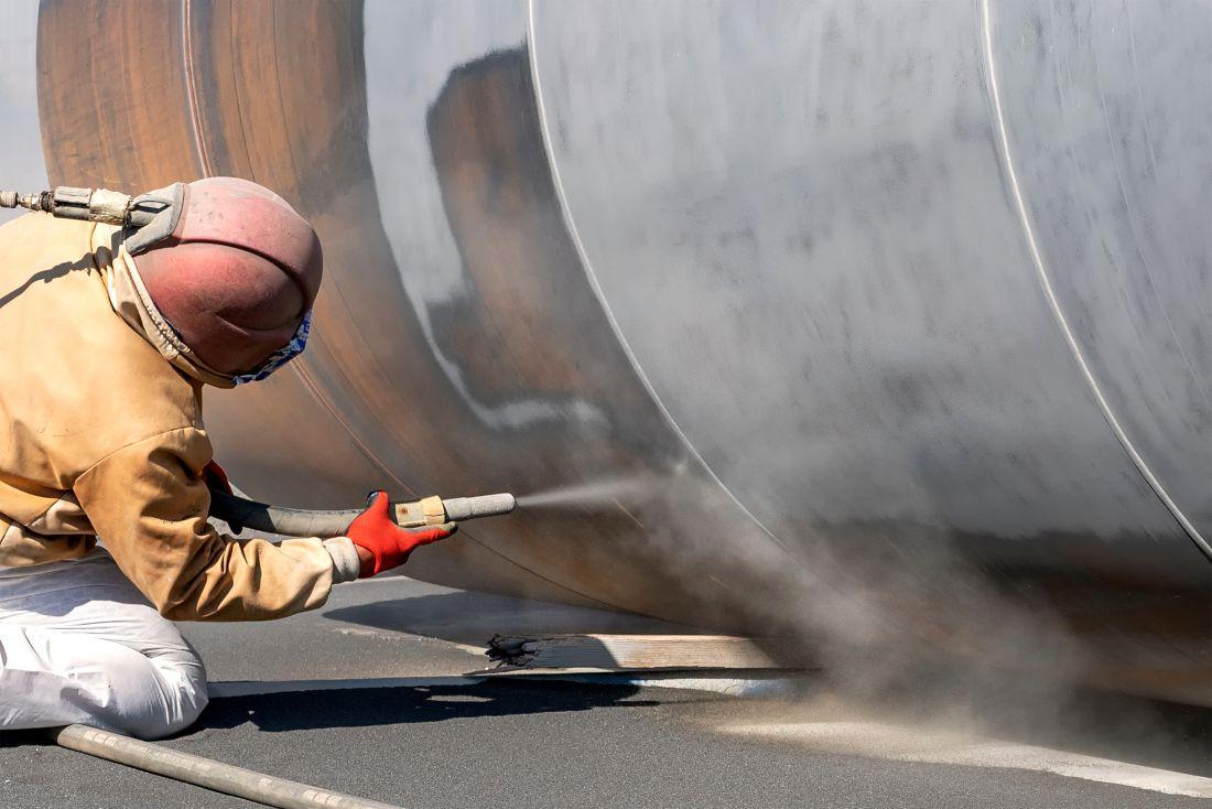 Arbeiter im Schutzanzug strahlt große Metalloberfläche mit Sandstrahlschlauch ab