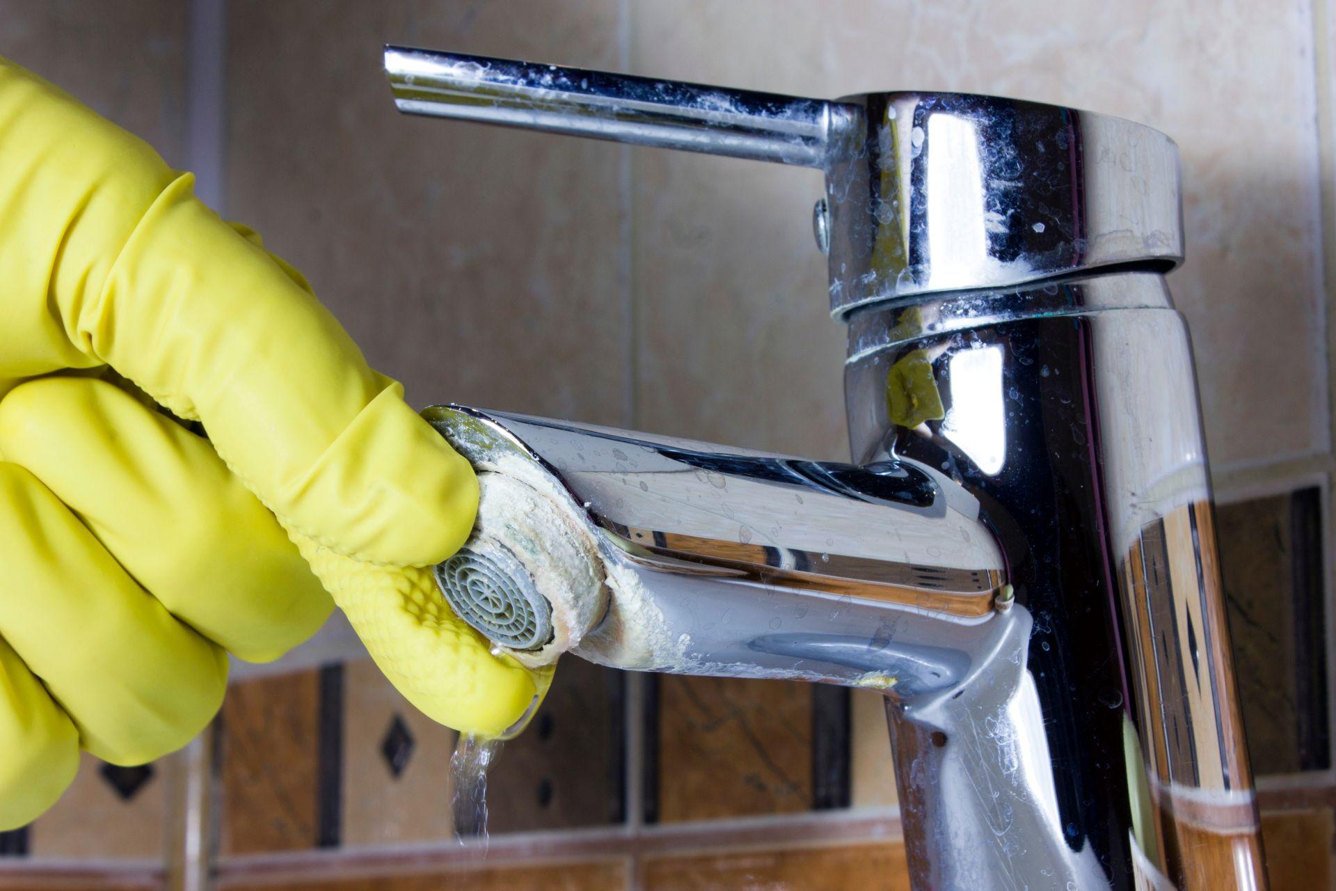 Hand mit gelbem Reinigungshandschuh entfernt Kalk von einem verkalkten Wasserhahn – haeufige Folge von hartem Leitungswasser ohne Enthaertun