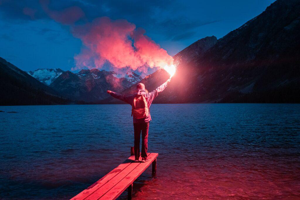 Person steht auf einem Steg vor Bergkulisse und setzt Leuchtmunition als starkes Lichtsignal in der Dunkelheit ein